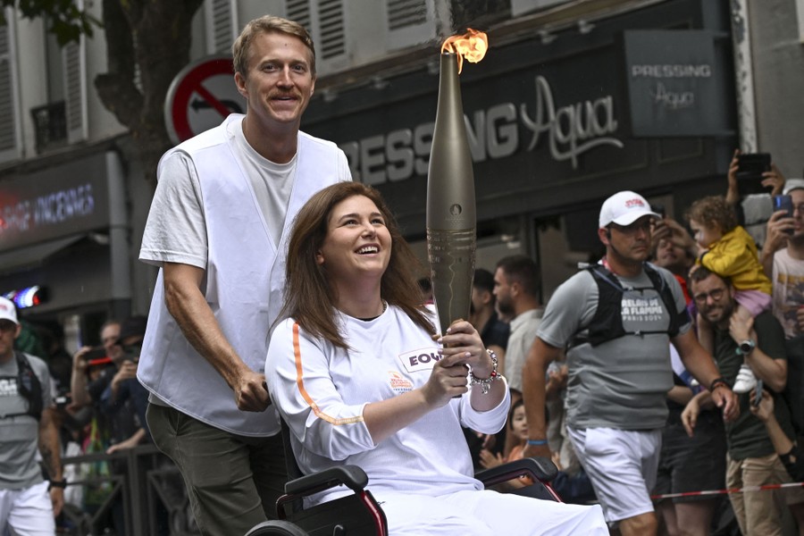 A person pushes another person in a wheelchair, who holds an Olympic torch.
