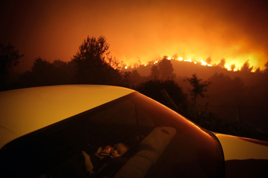 A child sits inside a car as a forest fire blazes in the distance.