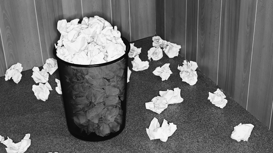 black and white photo of a trash bin filled with and surrounded by crumpled up wads of paper
