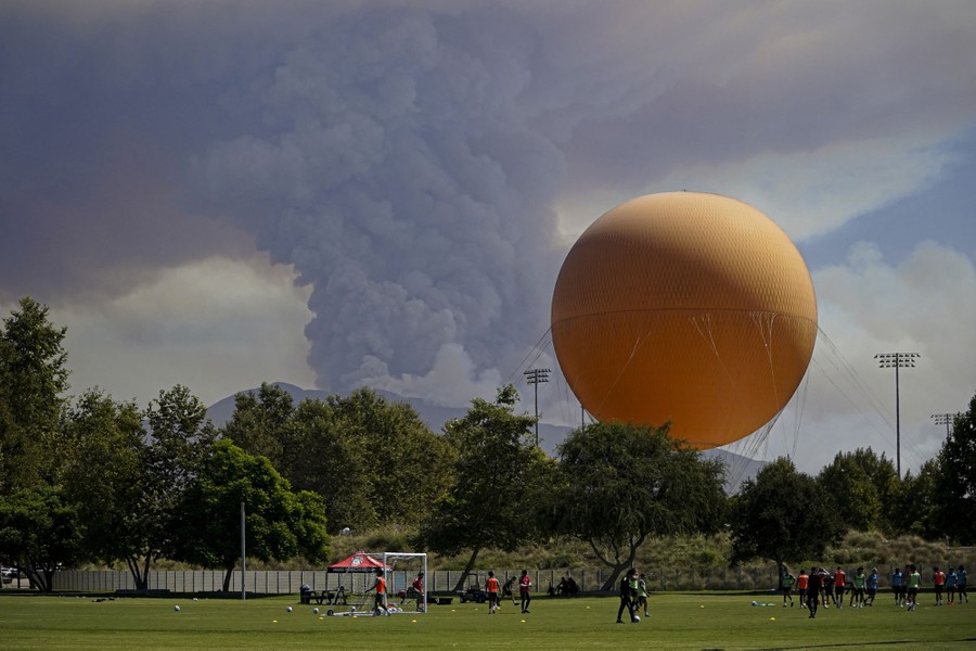 A plume of smoke created by a wildfire, seen beyond a group playing soccer and a large inflated balloon