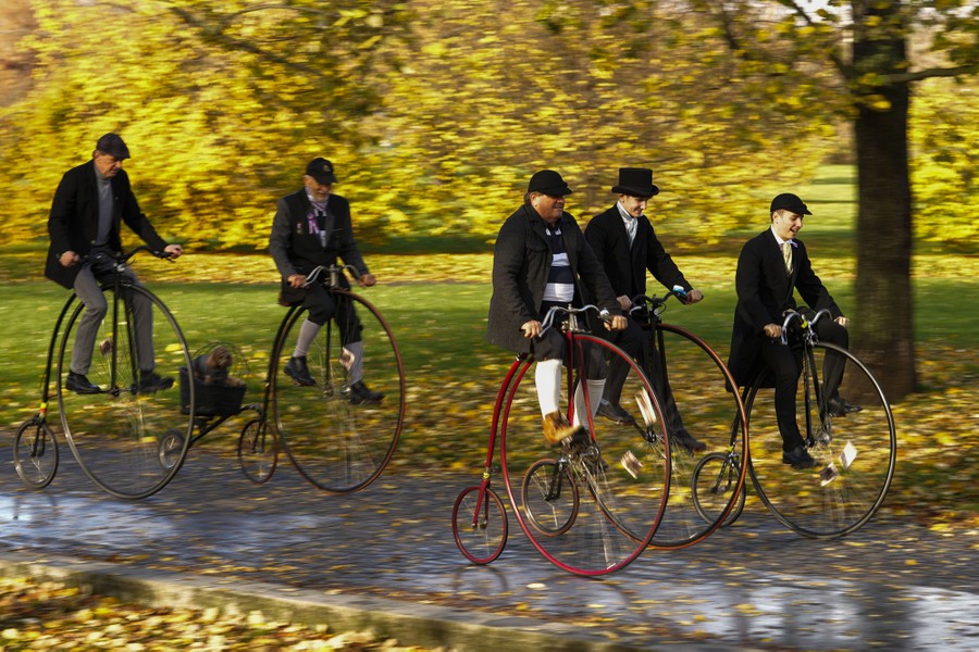Five people (and one dog) ride old-fashioned bicycles with large front wheels through a park.