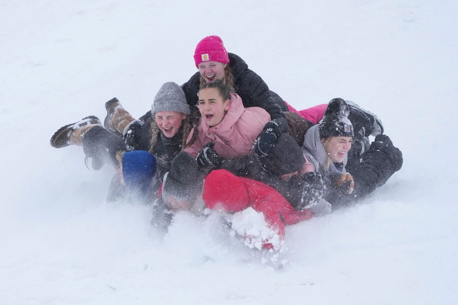 A pile of about eight people in winter gear laugh as they sled down a snowy hill.