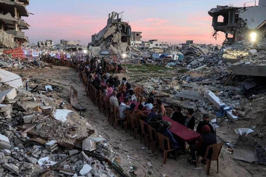 Many people sit on either side of a very long table, set up along a pathway through many destroyed buildings.