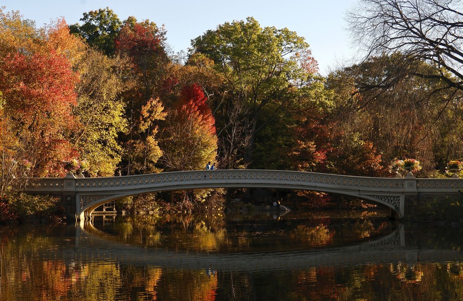 Bright fall foliage is reflected in a pond as people walk across a bridge.