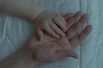 An image of an infant's hand on top of an adult's hand, both resting on a white comforter