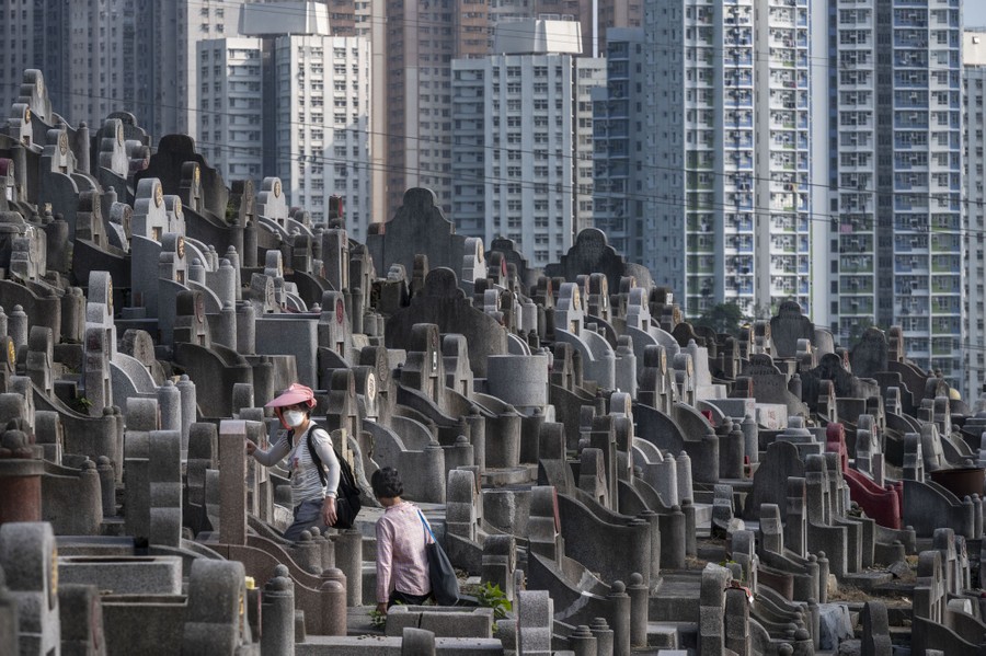 People visit a gravesite in a hillside cemetery beside skyscrapers.