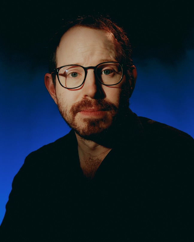 Ari Aster wearing glasses against a blue background
