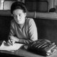 Simone de Beauvoir sits at a table at the Café de Flore, looking into the distance. She has pieces of paper in front of her and holds a pen. Her purse is on the table beside her.
