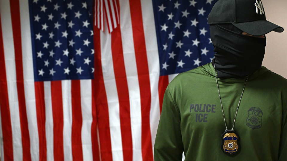 An image of a person wearing a shirt that reads "Police ICE" and who has their face fully covered with a neck gaiter and head covered with a Yankees baseball cap, standing in front of an American flag.