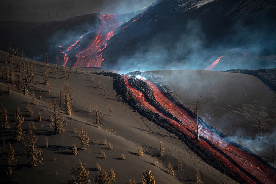 Lava flows down a series of mountain slopes, causing one tree to smolder.