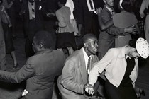 A black and white photo of two men and a woman dancing, on a crowded dance floor