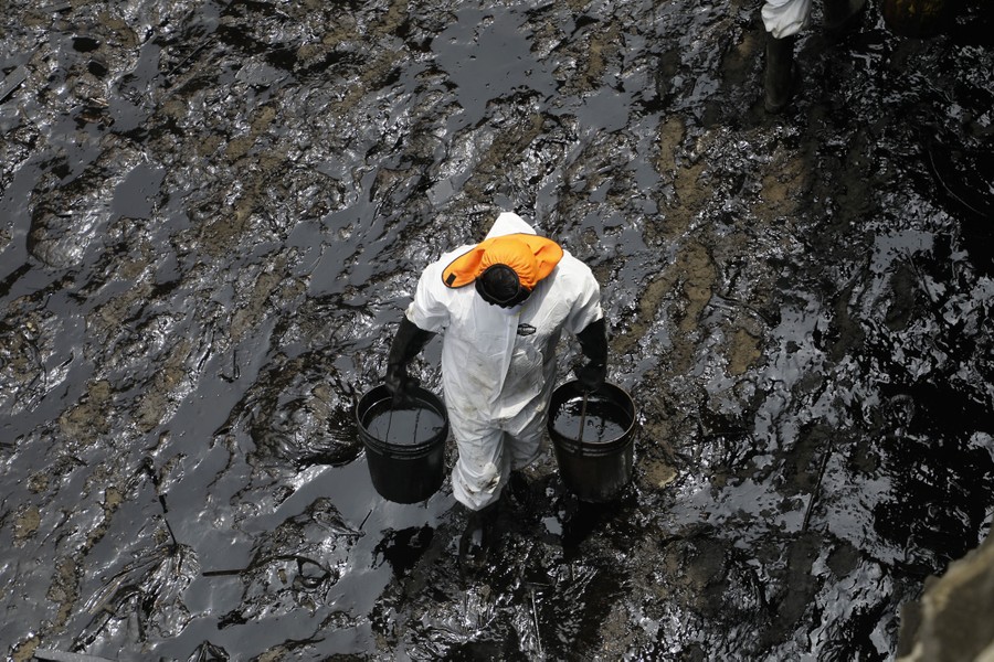 A cleanup worker carries two large buckets filled with spilled oil, on a rocky shoreline.