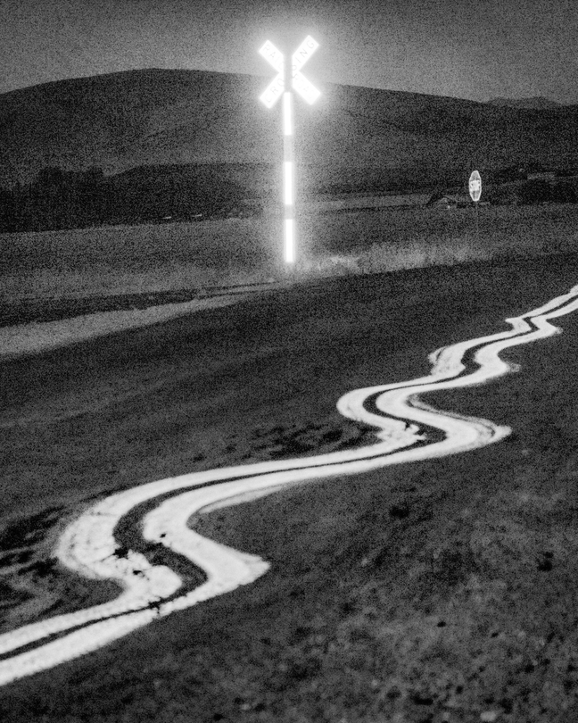 A black-and-white photo of a glowing white traffic sign and a swirly white line on the ground, with a hill in the background