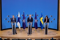 Three people stand at lecterns on a stage, with the flags of Finland, Ukraine, and NATO between them, during a press conference in Brussels.