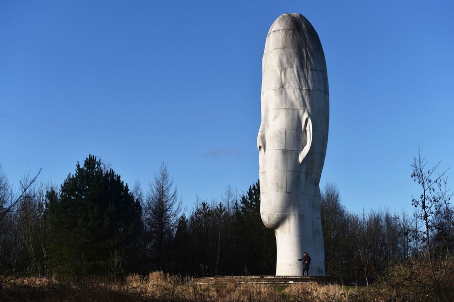 A person stands beside an enormous statue of a girl's vertically stretched head.