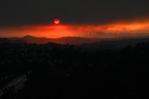 An aerial image showing vehicles driving on a freeway as the sun sets behind smoke from wildfires in Los Angeles