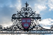 The Harvard seal adorns the top of a gate that reads "Soldiers Field Harvard University," with a partly-cloudy blue sky in the background.