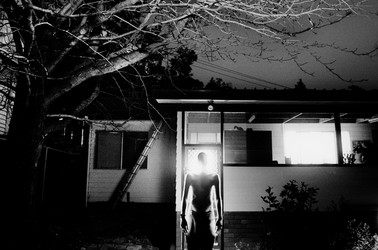 a man standing in darkness before a house, glowing from within