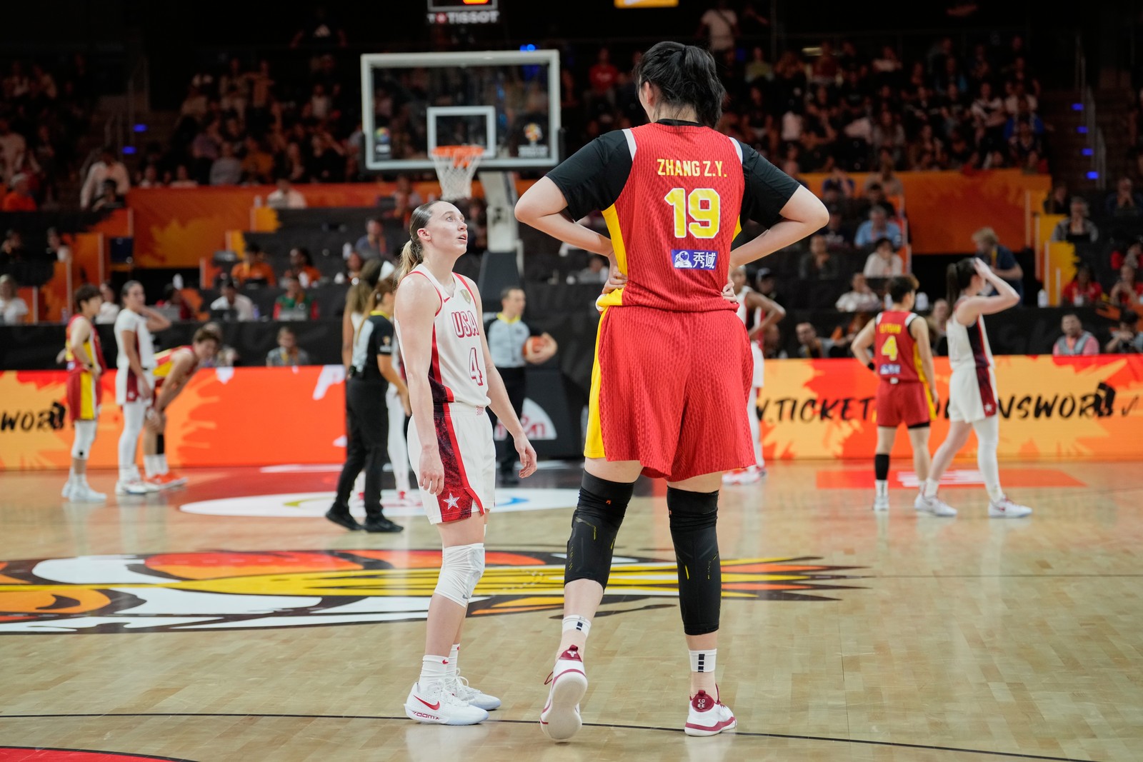 Two basketball players from opposing teams talk on a court. One looks up at their much-taller opponent.