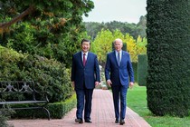 Xi Jinping and Joe Biden, both wearing blue suits, walking side by side on a brick path through a garden