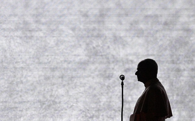 Photograph of Pope Leo’s black silhouette from the side as he speaks in front of a microphone.