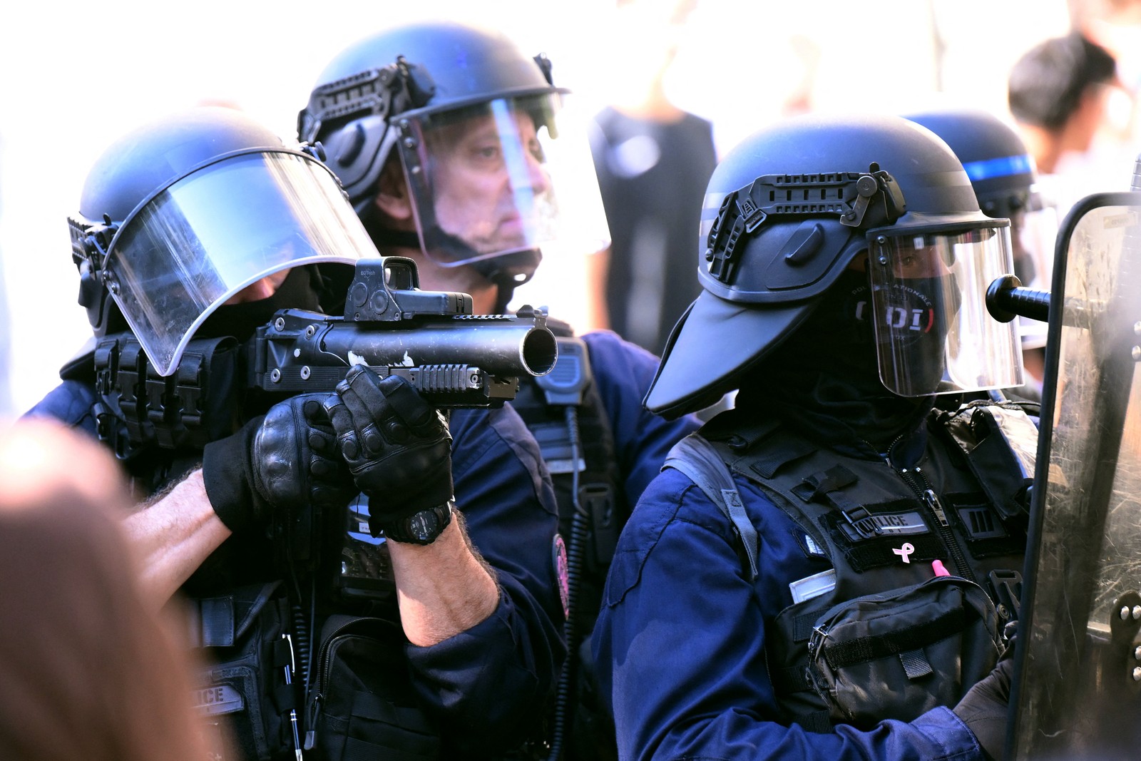 A police officer, one of three officers in riot gear, aims a riot-control weapon.