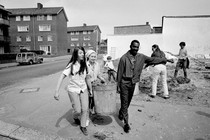 Image of two young women and one young man carrying a trash can together down a neighborhood sidewalk.