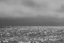 A black-and-white photograph of ocean waves below a stormy sky