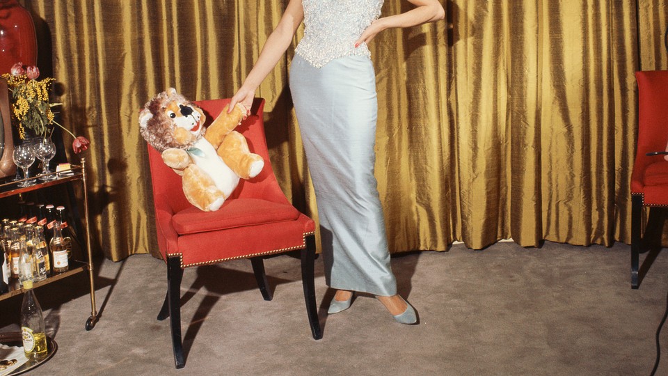 Vintage photo of a woman in an evening gown standing in front of a gold curtain, dangling a stuffed lion from her hand