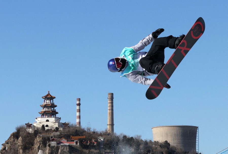 A snowboarder is seen in mid-air, during a jump, in front of distant smokestacks and a pagoda.
