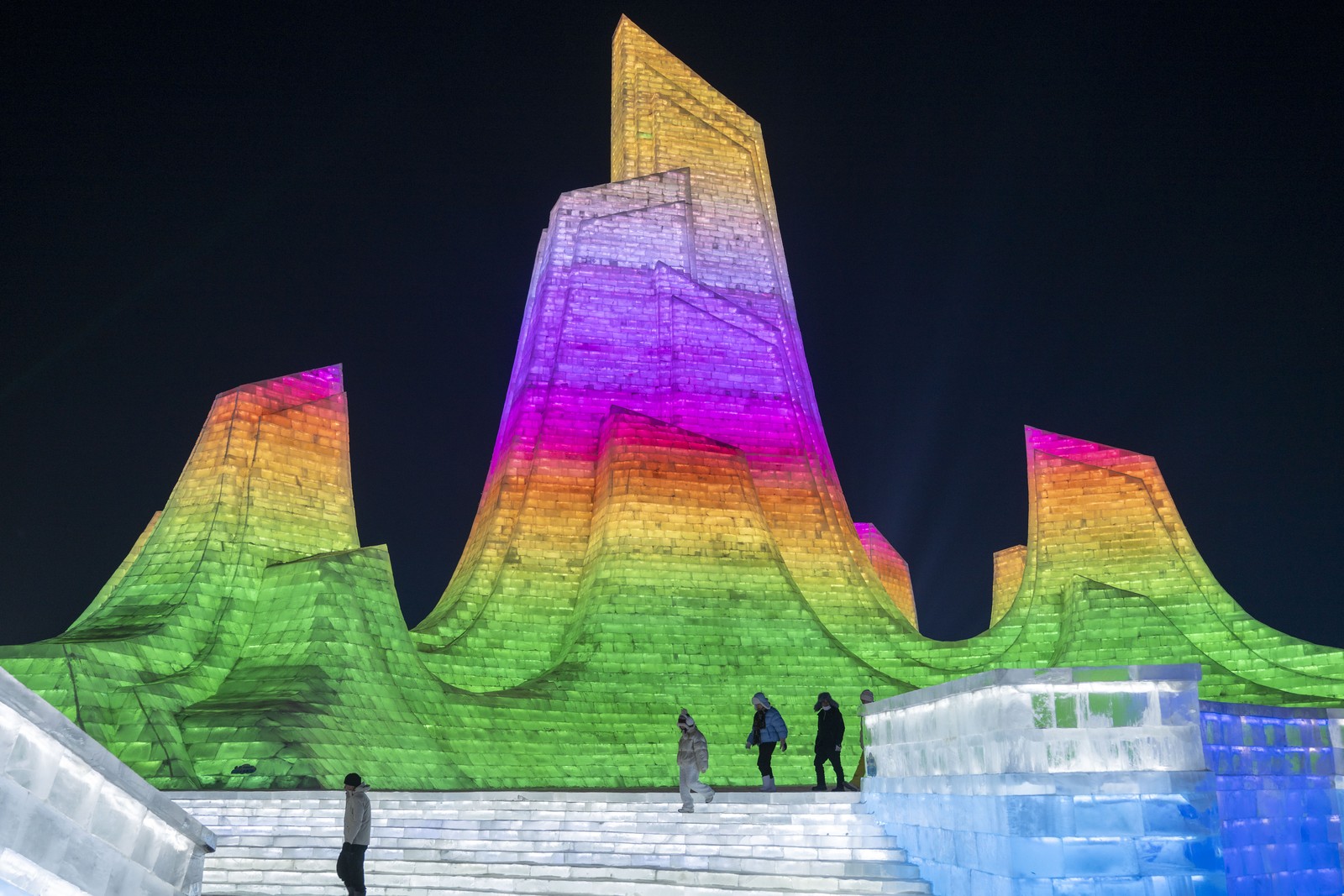People walk past tall ice sculptures that are lit in a rainbow of colors.