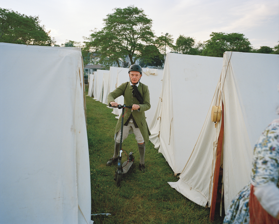 photo of reenactor wearing period dress wearing a modern helmet and riding an electric scooter along the grass between two rows of white canvas tents