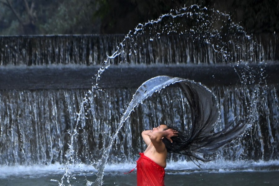 A woman tosses her long wet hair back, throwing water in an arc above her, while standing in a river near a manmade waterfall.