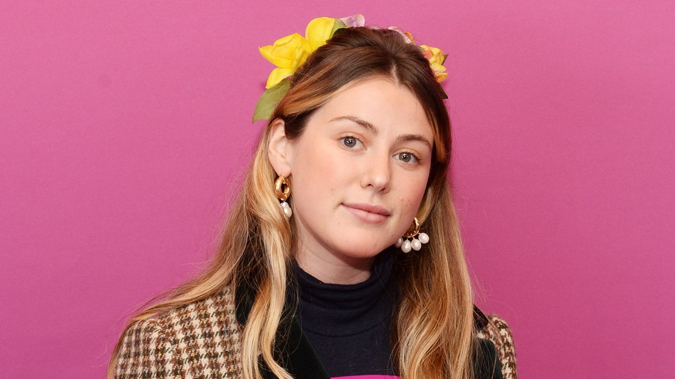 Caroline Calloway stands in front of a pink backdrop looking straight into the camera.