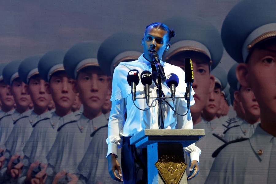 A performer stands at a podium with several microphones mounted on it, while a video display shows images of many uniformed people in the background.