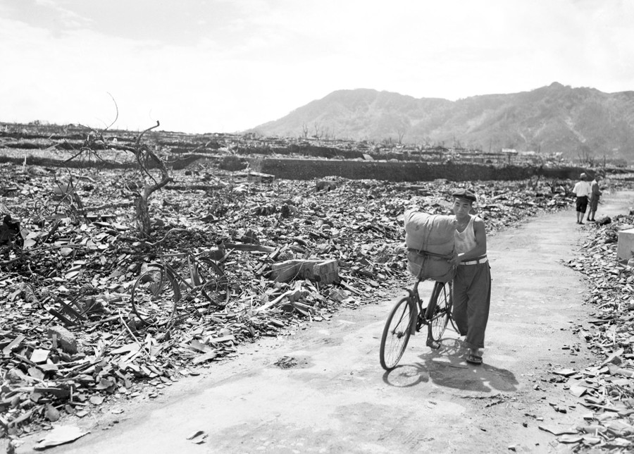 A civilian pushes a bicycle, carrying a bundle, on a cleared path through a destroyed city, following the atomic bombing of Nagasaki, Japan.
