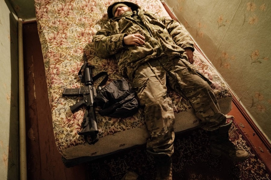A soldier rests on a bed, in full gear, with his weapon beside him.