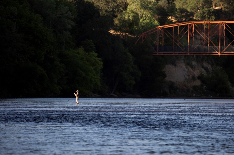 A person fishes while standing in the middle of a shallow river.