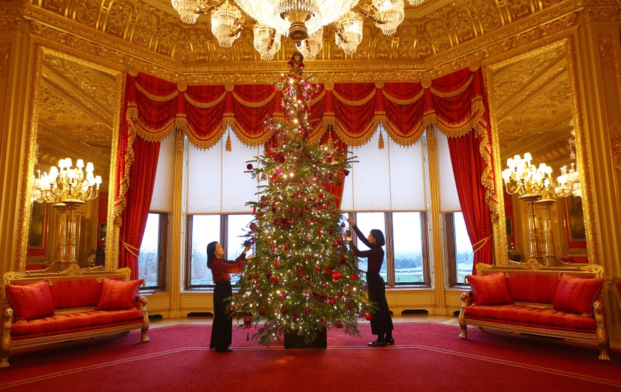 Two people adjust ornaments on a tall Christmas tree inside an ornate room of Windsor Castle.