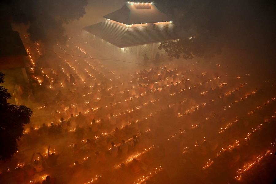 Hundreds of Hindus sit in rows among thousands of lit candles and smoke, outside a temple, at night.