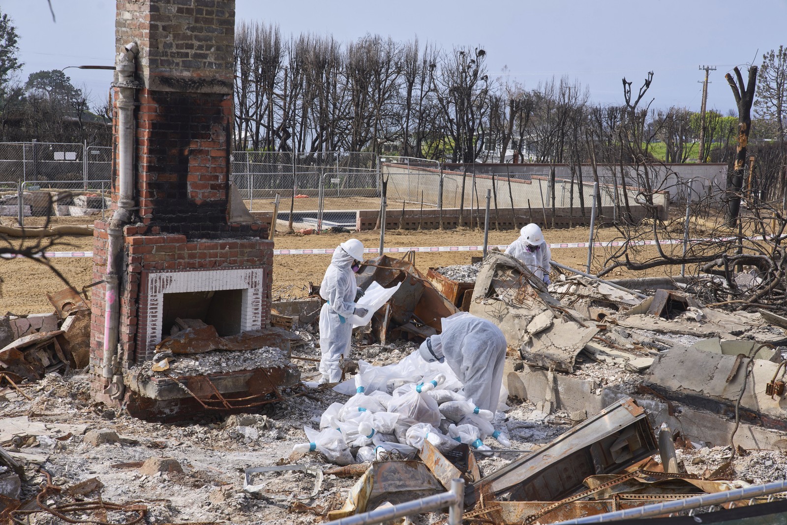 Workers in protective gear remove hazardous materials from the site of a burned house.