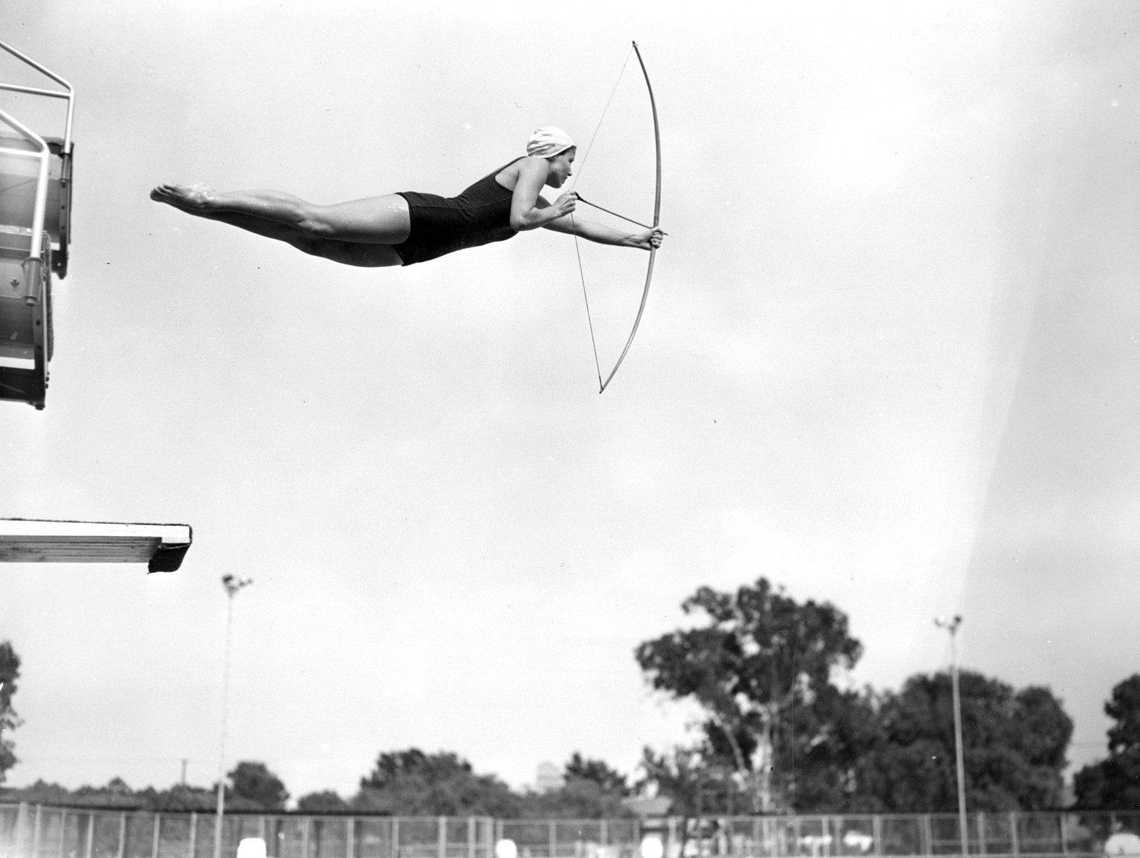 A woman is seen in mid-air during a dive into a pool, holding a bow and drawing back an arrow.