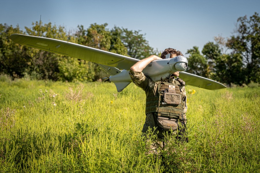 A soldier carries a fixed-wing surveillance drone on his shoulder in a field.