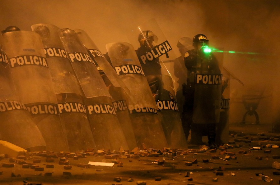 A group of riot police stand behind their shields; one of the officers seems to have a green laser aimed at him.