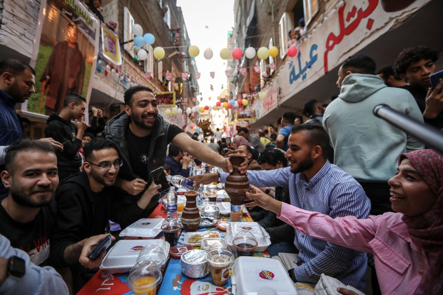 Ένα μεγάλο πλήθος κάθεται σε ένα μακρύ τραπέζι σε έναν δρόμο, απολαμβάνοντας ένα γεύμα.