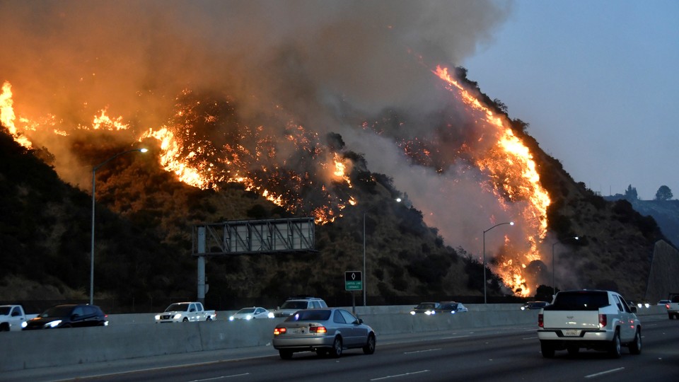 A fire burns on a hillside next to a freeway as cars drive by.