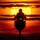 A man rides over a hill on a motorcycle as the sun rises in the background.
