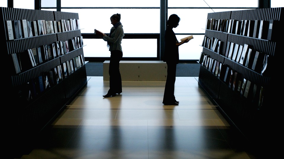 Two women stand next to rows of books, reading. 