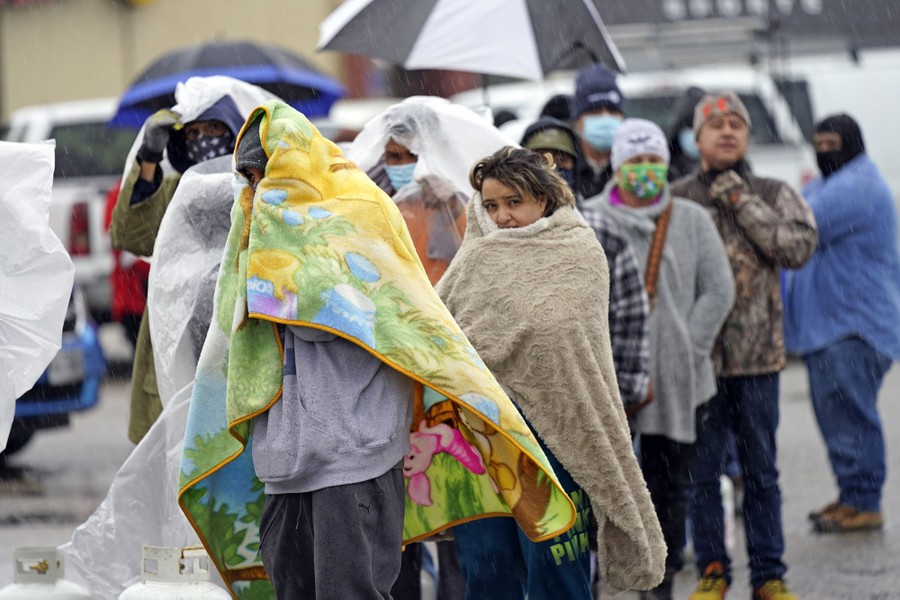 People stand in line in a parking lot, wrapped in blankets and plastic sheets.