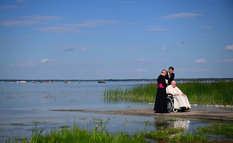 Two people attend to Pope Francis, who is in a wheelchair, on the shore of a broad lake.